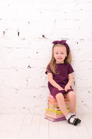 blond girl sitting on a pile of books against a white brick wallの写真素材