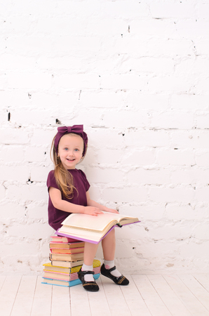 Girl reading a book, sitting on a pile of books, against a white brick wallの写真素材