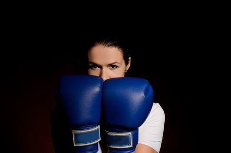 Girl in boxing gloves covers face protection pose against a dark backgroundの写真素材
