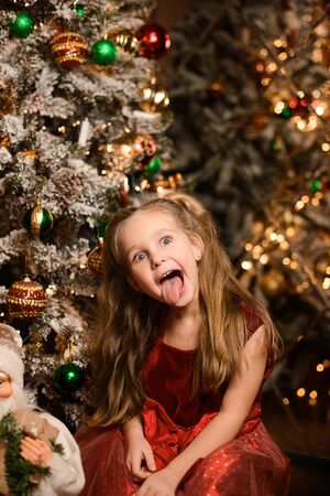 Portrait of beautiful girl near festive Christmas tree trimmed with balls, against background of mirror, bokeh from lights of garlandsの写真素材