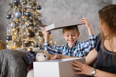 Boy was delighted and surprised at gift he saw in big gray box, in his mother's hands, sitting on sofa against background of Christmas tree glowing with them bokehの写真素材