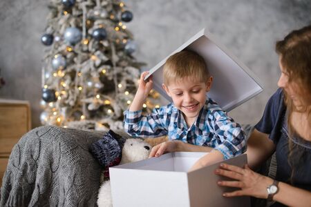 Boy was delighted and surprised at gift he saw in big gray box, in his mother's hands, sitting on sofa against background of Christmas tree glowing with them bokehの写真素材