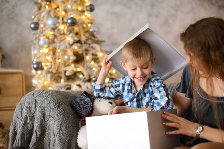 Boy was delighted and surprised at gift he saw in big gray box, in his mother's hands, sitting on sofa against background of Christmas tree glowing with them bokehの写真素材