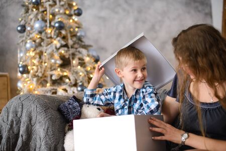 Boy was delighted and surprised at gift he saw in big gray box, in his mother's hands, sitting on sofa against background of Christmas tree glowing with them bokehの写真素材