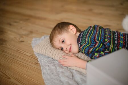 boy in sweater lies on wooden floor, his head on knitted pillow, and smilesの写真素材