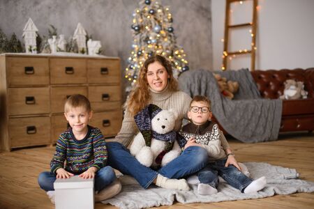 Mom with two sons in sweaters, hugging, sitting on wooden floor, in room with Christmas tree, next to toy bearの写真素材