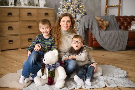 Mom with two sons in sweaters, hugging, sitting on wooden floor, in room with Christmas tree, next to toy bearの写真素材