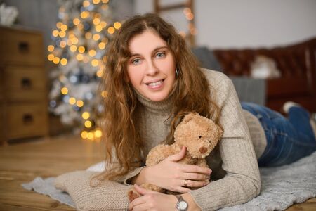 Young happiness woman with curly hair in an embrace with toy teddy bear, lies on wooden floor, against the background of Christmas treeの写真素材