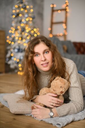 Young happiness woman with curly hair in an embrace with toy teddy bear, lies on wooden floor, against the background of Christmas treeの写真素材