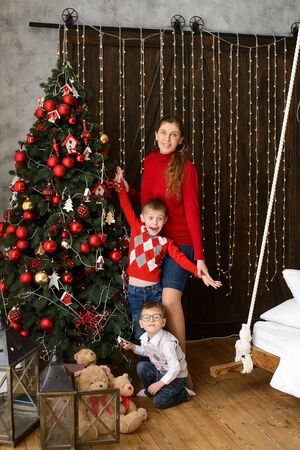 Mom with little sons decorates Christmas tree with red balls, against background of wooden wall with lights garlandsの写真素材