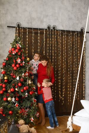 Mom with little sons decorates Christmas tree with red balls, against background of wooden wall with lights garlandsの写真素材