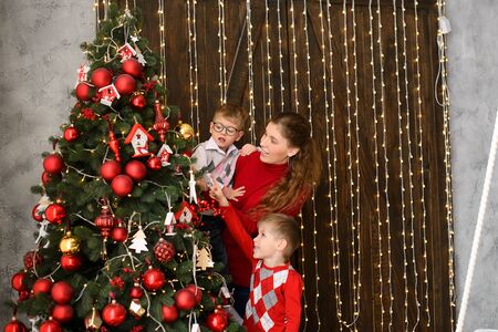 Mom with little sons decorates Christmas tree with red balls, against background of wooden wall with lights garlandsの写真素材