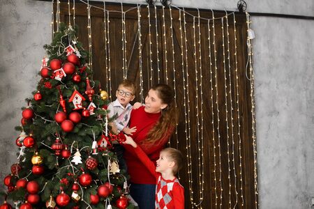 Mom with little sons decorates Christmas tree with red balls, against background of wooden wall with lights garlandsの写真素材