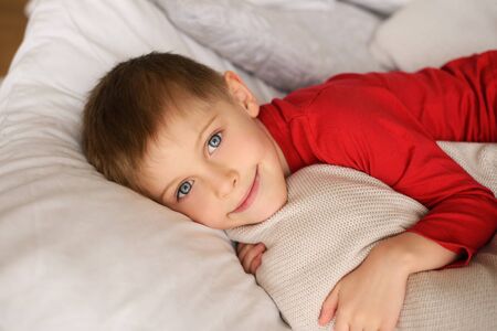 handsome light-eyed boy in red jumper lies among white pillows and blanketsの写真素材