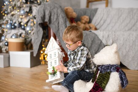 boy with glasses sitting on wooden floor plays with white toy house on background of Christmas tree and gift boxes.の写真素材