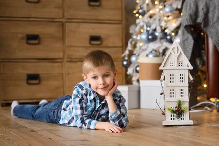 boy lies on wooden floor, near toy house, against background of Christmas tree with gift boxes underneathの写真素材