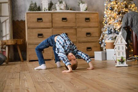 boy stands in an acrobatic pose on wooden floor, against background of Christmas tree, glowing with lights and bokehの写真素材