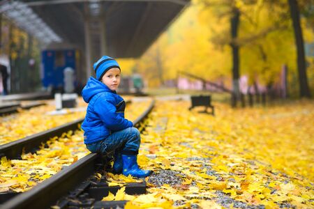 boy in blue hat, jacket, boots, sits on rails against background of train, and in autumn park with yellow fallen leavesの写真素材