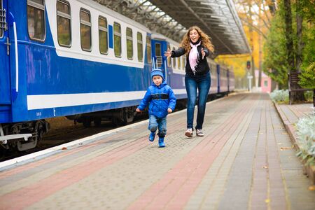 Mom catches up with laughing runaway little son along blue train at stationの写真素材