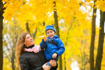Mom with son in her arms walks in autumn park among maple trees with yellow foliageの写真素材