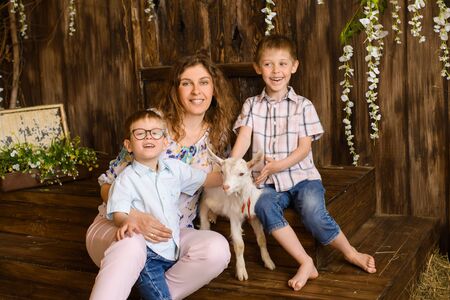 sitting young mother and two her sons kids with little white goat on dark wooden background with flowers and hayの写真素材