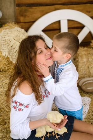 son kisses his mother on cheek, dressed in Ukrainian embroidered shirts, sit in hayloft, play with live chickens on Easter holidaysの写真素材