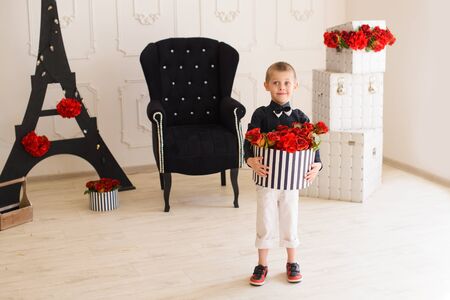 Beautiful little son in dark shirt and bow tie with gift box in his hands full of red rosesの写真素材
