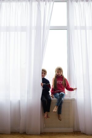 brother and sister are sitting on windowsill, near window glass, they want to walk on street, but in world there is  coronavirus pandemic, and they remain at home in quarantineの写真素材
