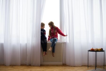 brother and sister are sitting on windowsill, near window glass, they want to walk on street, but in world there is  coronavirus pandemic, and they remain at home in quarantineの写真素材