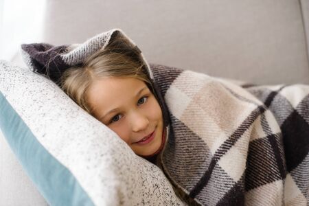 Cute girl lies on sofa and pillow, hiding in cozy blanket, daytime sleep in roomの写真素材