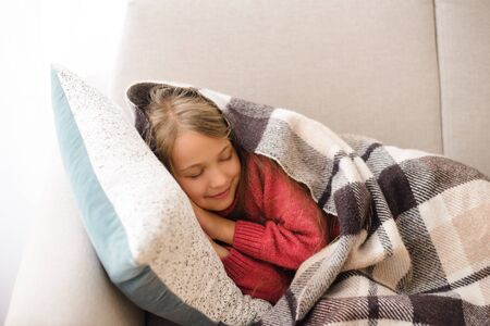 Cute girl lies on sofa and pillow, hiding in cozy blanket, daytime sleep in roomの写真素材