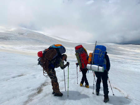 Three tired climbers with backpacks and trekking poles stand on the slopes of Elbrus. Snowy slopes of the northern Elbrus region. Mountain winter landscape.の写真素材