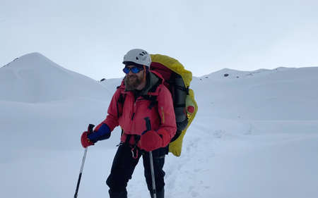 Smiling climber in a helmet, sunglasses, a backpack and trekking poles walks along a snow-covered...