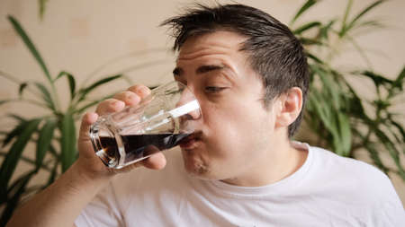 A charismatic guy drinks soda from a glass glass. Close-up. Green plants on the background.の写真素材