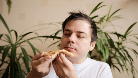 The young man takes a slice of pizza, takes a bite with passion and chews it. The guy looks at a slice of pizza adoringly. Delicious cheesy pizza.の写真素材