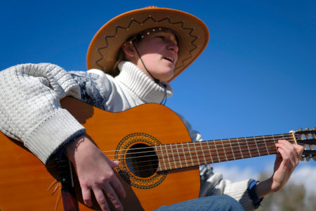 Portrait of a female guitarist singing songs with a guitar against a blue sky background. A musician in a cowboy hat and a white sweater plays the guitar, focusing on the hand and strings. The concept of relaxation and leisure when a guitarist sits outside and enjoys musicの写真素材