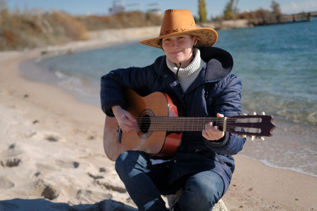 A young woman is sitting on the beach with a guitar in her hands. She's wearing a brown cowboy hat and a blue jacket. The guitarist plays on the shore of a blue lake on a sunny day.の写真素材