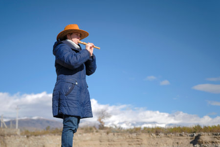 A cowboy woman in a wide-brimmed hat and a blue coat is playing a flute in a field. The sky is blue, the clouds are white, and mountains are visible in the background. A piper's concert in nature. The musician plays the fluteの写真素材