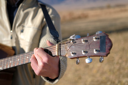 Close-up focus on a guy's hand clenching a chord on the neck of a guitar. A man is playing guitar in a field. The guitar is a vintage model with a black strap. The musician is wearing a white jacket and enjoying playing the guitarの写真素材