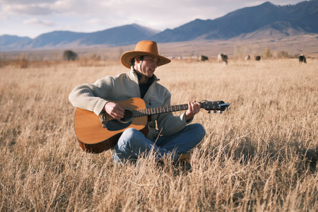 The concept of freedom. A young cowboy herds a herd of cows in an arid field in the mountains. A musician in a cowboy hat is sitting in a field in the dry grass and playing the guitar. The guitarist is enjoying his village life.の写真素材
