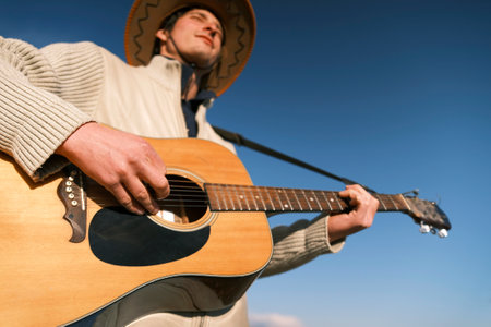 A cowboy plays a guitar against a blue sky, a view from below. The focus is on the hand and the strings. A man in a cowboy hat is playing his favorite tune on the guitar with his eyes closed. Guitar lessons. The soul of a musicianの写真素材