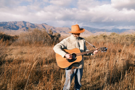 A cowboy with a guitar in his hands. A man in a cowboy hat plays guitar in an arid field on a warm autumn day in the mountains. The image is made in a peaceful and relaxed style, as the guitarist is surrounded by nature and enjoys himselfの写真素材