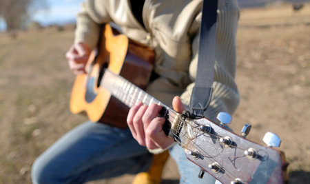 A man is playing guitar in a field. Focus on the guitar neck, strings and arm. The guitar is old and worn. Guitar lessons in the fresh air. The scene is laid-back and casual.の写真素材