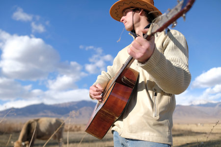 A man in a cowboy hat and a blade of grass in his mouth is playing guitar in a field against a background of blue sky, white clouds and mountains. An authentic shepherd musician herds a herd of cows. Rural life on the ranch.の写真素材