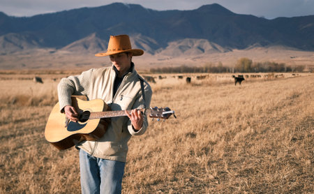 An authentic cowboy with a guitar is dancing in an arid field in the mountains. A man in a cowboy...