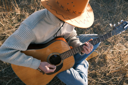 A cowboy is sitting in a field with a guitar in his hands, top view. A man in a cowboy hat plays guitar in an arid field. The concept of freedom and relaxation when a musician enjoys time spent outdoorsの写真素材