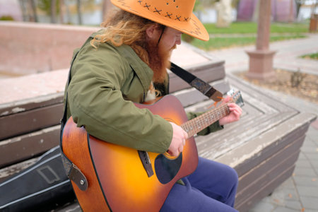 A man with long wavy red hair and beard, wearing a cowboy hat, is playing guitar while sitting on a park bench. An authentic portrait of the guitarist. The musician composes a new melody on the guitar.の写真素材