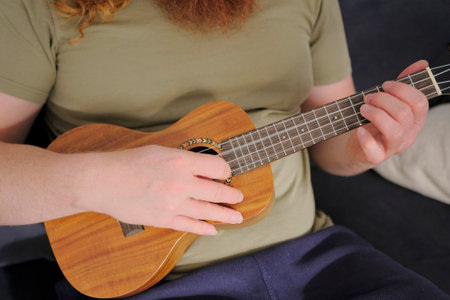 Ukulele singer. A man plays the ukulele, close-up, focus on the hand and strings. The musician learns a new melody. The guitar is small and made of wood. The guitarist is sitting on the couchの写真素材