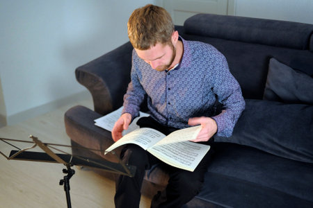 A man is sitting on the couch, with sheet music in front of him. The musician reads the notes and focuses on them. A student is rehearsing or preparing for a performance while sitting on the couch at homeの写真素材