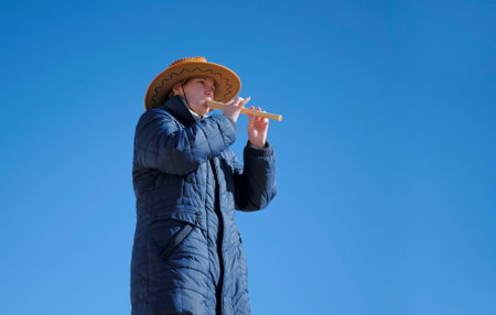 A young female student in a cowboy hat and a blue coat plays a flute against a blue cloudless sky. The piper gives a concert in the fresh air. A musician learns to play the trumpet. Summerの写真素材